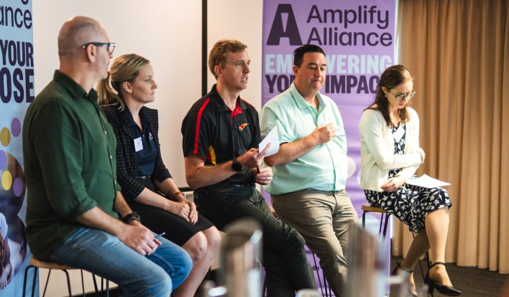 Five panelists sit on stools, left to right, in a brightly lit room with a purple banner backdrop; they wear varied attire, holding papers, creating a professional, engaged, and informative atmosphere.