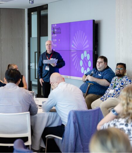 A panel discussion unfolds in a brightly lit room, featuring three speakers before an audience seated around a table; a large screen displays a purple logo, creating a modern, professional ambiance.