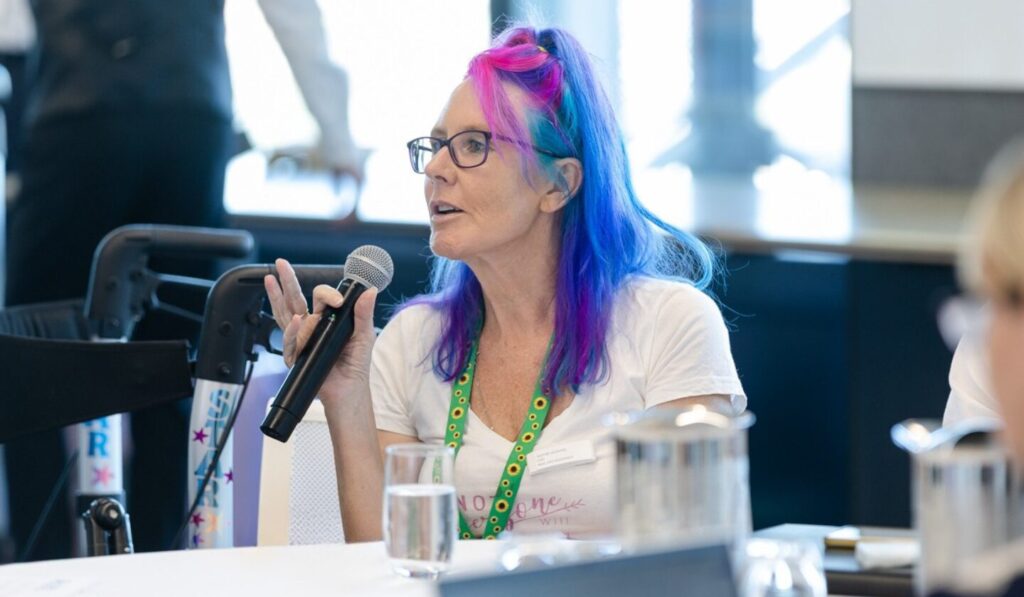 A woman with vibrant blue and pink hair speaks into a microphone at a table, wearing glasses and a white shirt with a sunflower lanyard; bright, natural light illuminates the scene.