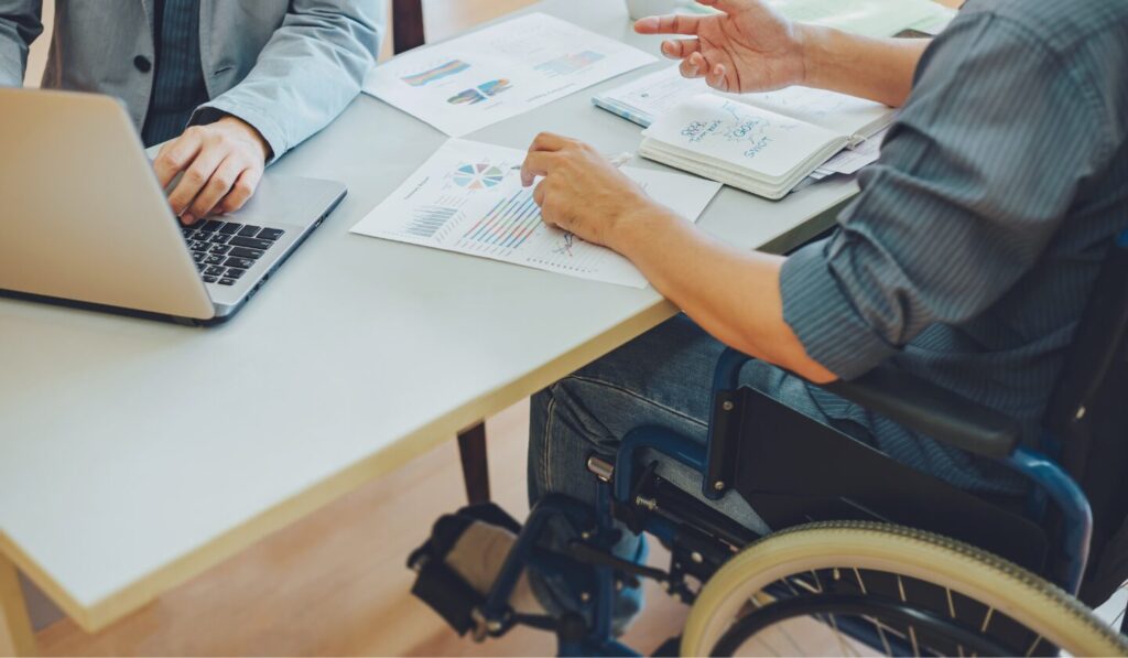 Two businesspeople collaborate at a bright, modern table; one uses a laptop, the other, in a wheelchair, gestures at charts and a notebook with "GOAL" written on it.