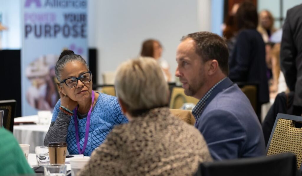 A woman with glasses, in blue, sits at a round table with a man in a blue jacket and a woman in a patterned top, engaged in conversation under bright, neutral lighting.